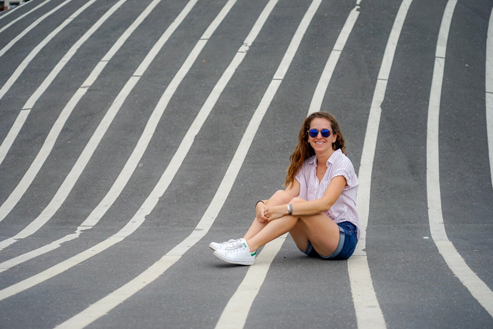 Young woman in summer clothes and sunglasses sitting on the pavement floor and smiling.
