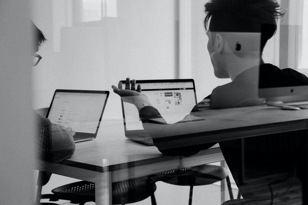 A black and white picture of a man looking at his laptop and explaining something to another person.